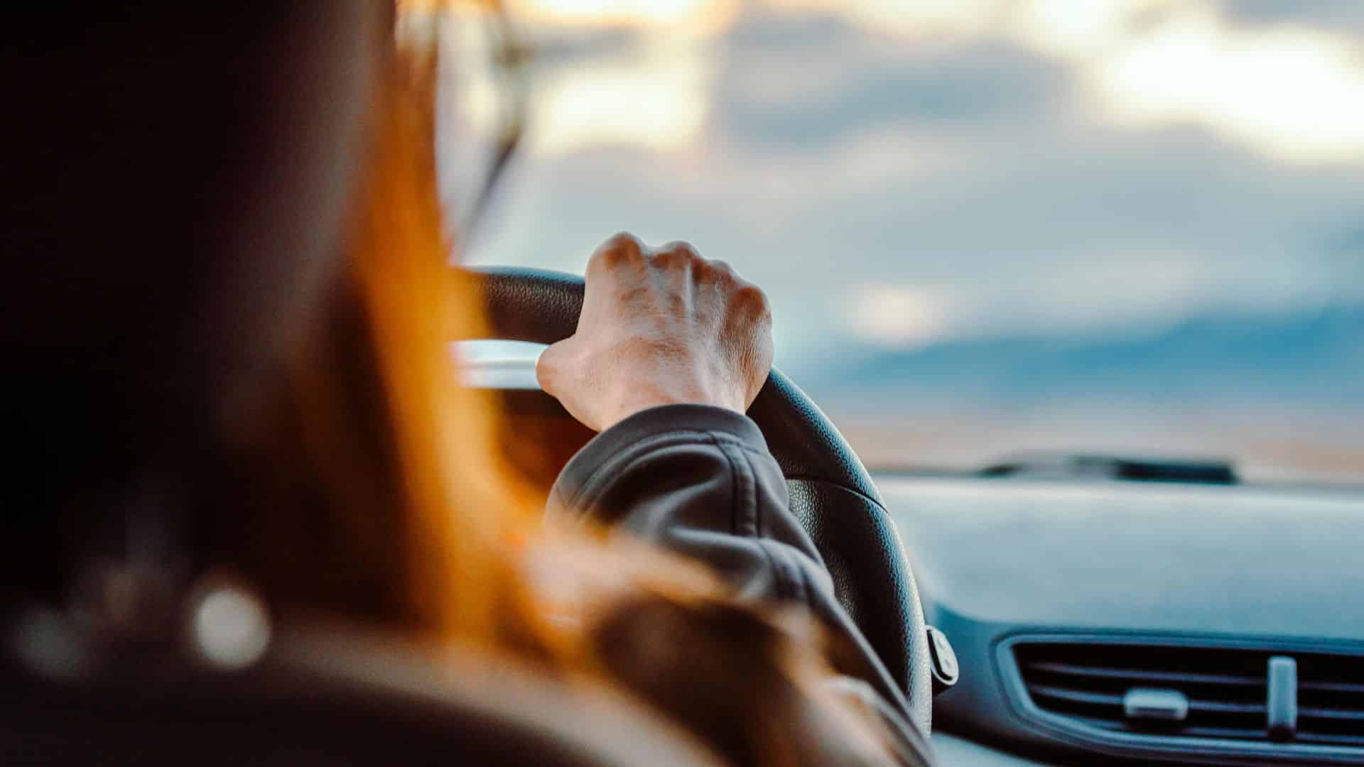 Conductrice vue de dos, une main sur le volant et la route en arrière-plan.